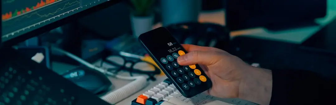 Hand using a phone calculator at a desk below monitors showing charts in a dark office