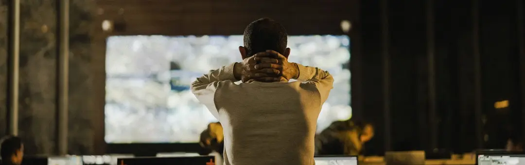 Man with hands behind his head facing a large video wall in a dark control room