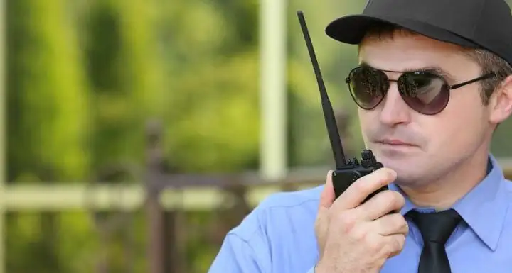 Security guard in sunglasses and a cap speaking into a two-way radio outdoors