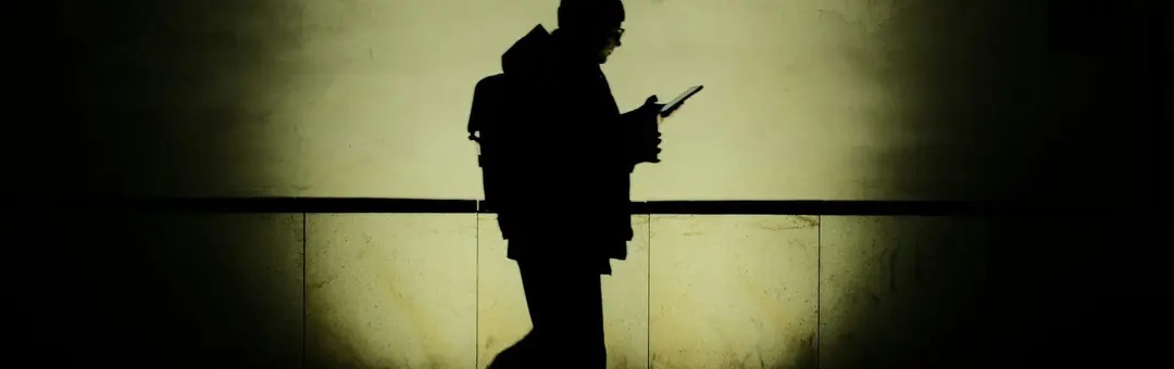 Silhouette of a person with a backpack using a phone against a glowing wall at night